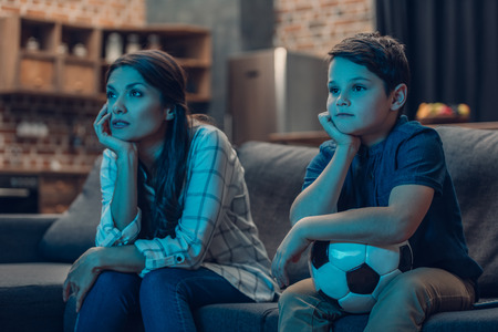 Bored little boy and his young mother watching a soccer game on couch in living roomの写真素材