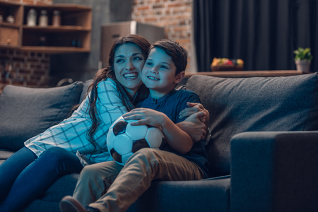 Little boy and his beautiful mother hugging on couch while watching a soccer gameの写真素材