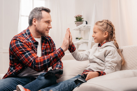 smiling father and daughter giving high five while playing with joysticks at homeの写真素材