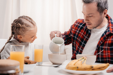 smiling little girl looking at father pouring milk into bowl with flakes at breakfastの写真素材