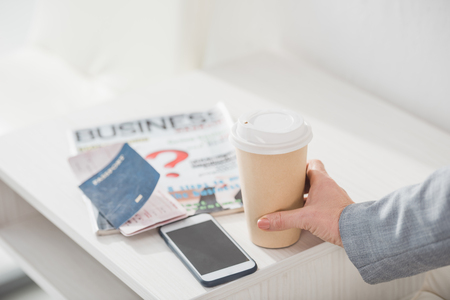 cropped shot of businesswoman holding paper cup of coffee on table with smartphone and flight ticketの写真素材