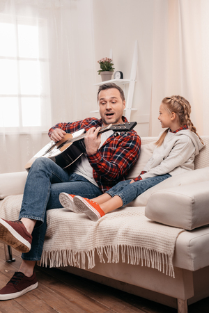 cute little girl looking at happy father playing guitar at homeの写真素材
