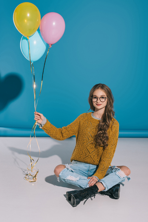 beautiful smiling teenage girl in eyeglasses holding balloons while sitting and looking at cameraの写真素材