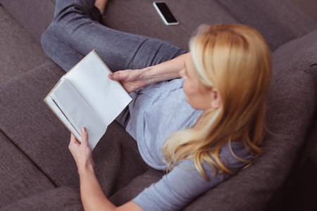 high angle view of blonde woman reading book while sitting on sofa at homeの写真素材