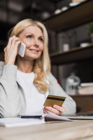 smiling blonde woman holding credit card and talking on smartphone at homeの写真素材