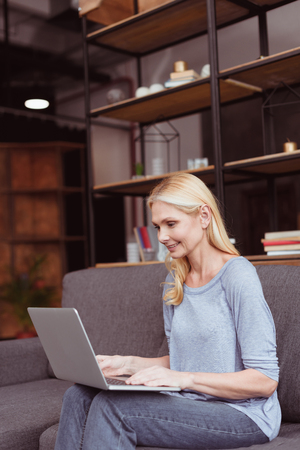 beautiful smiling blonde woman using laptop while sitting on sofa at home の写真素材