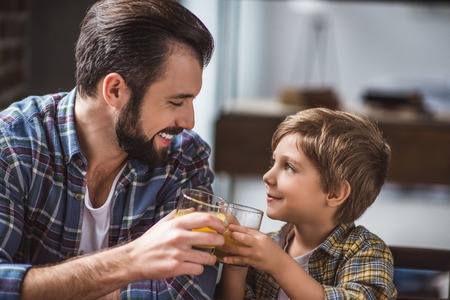 side view of smiling father and son clinking glasses of juice during breakfast at homeの写真素材