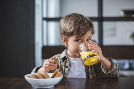 selective focus of cute little boy with cookie in hand drinking juice during breakfast at homeの写真素材
