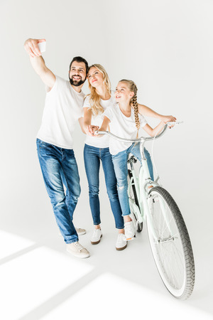 happy young family taking selfie with smartphone while child riding bicycle isolated on whiteの写真素材