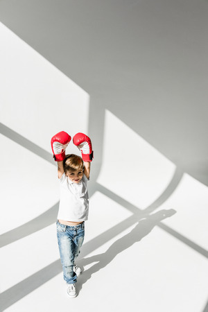 high angle view of cheerful little boy in boxing gloves raising hands on white の写真素材