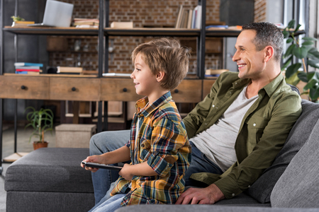 father and son watching tv at home while sitting on couchの写真素材
