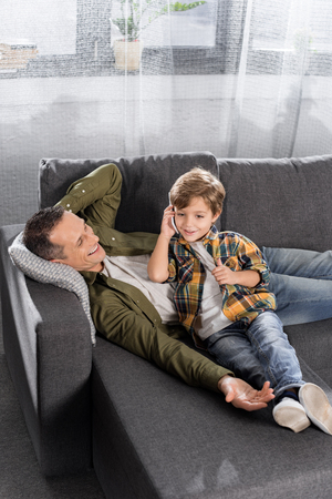 happy little son talking by phone while father laying on couchの写真素材