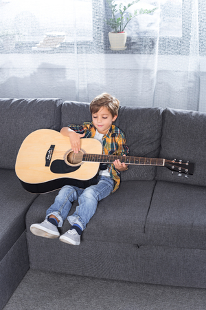 adorable little boy playing guitar on couch at homeの写真素材