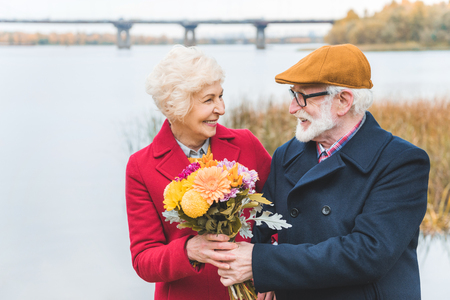 happy senior couple with bouquet of flowers standing at autumn lakeの写真素材