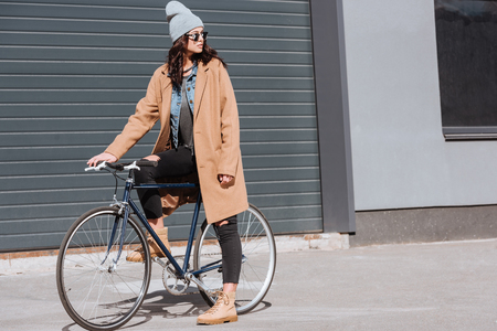 attractive woman in autumn outfit and black sunglasses sitting on bicycle and looking awayの写真素材
