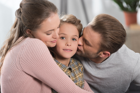 Two parents hugging and kissing little son while he is looking at cameraの写真素材