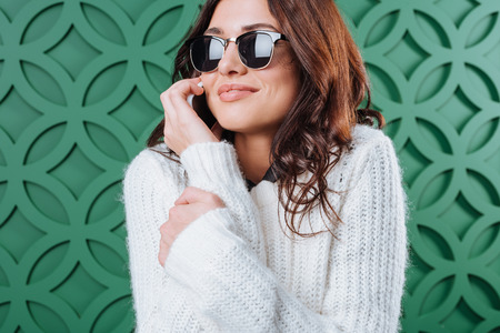 beautiful woman in white winter sweater and black sunglasses talking by smartphone on greenの写真素材