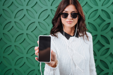 attractive woman in white winter sweater and black sunglasses listening to music and showing smartphoneの写真素材