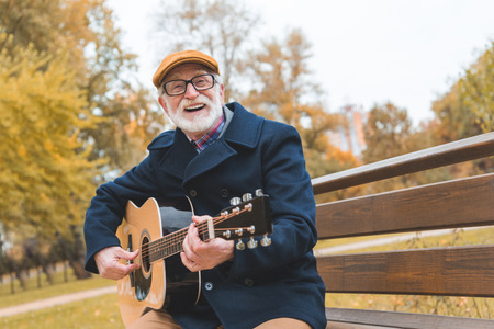 happy bearded stylish senior man playing on acoustic guitar in autumn park