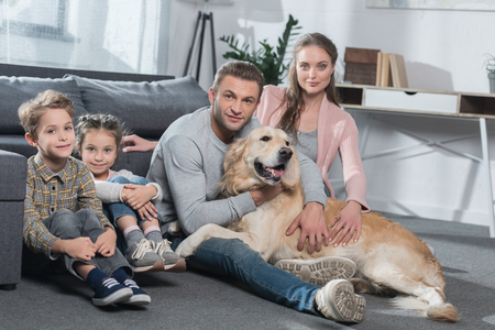 Family with two kids and dog sitting on floor in living room and looking at cameraの写真素材