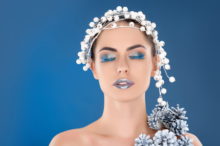portrait of beautiful woman with hair accessory, christmas pine cones, winter makeup and glitter, isolated on blueの写真素材
