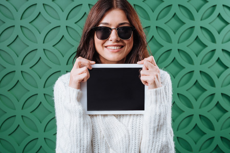 gorgeous woman in winter sweater and black sunglasses showing tablet computer and smiling の写真素材