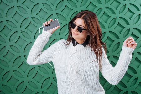 Stylish woman in white winter sweater and black sunglasses listening to music with smartphone and dancing on greenの写真素材