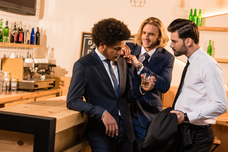 young businessmen looking at drunk friend standing with glass of whiskey in bar の写真素材