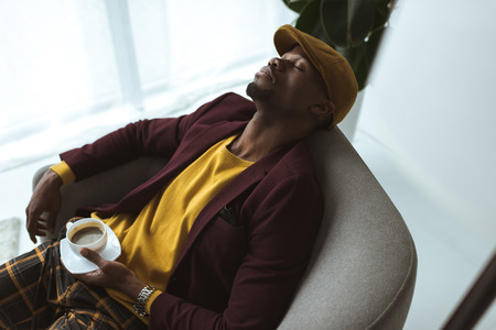 stylish tired african american man with coffee cup sleeping in armchair の写真素材