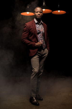 handsome fashionable african american man holding glass of cognac in dark smoky room with lampsの写真素材