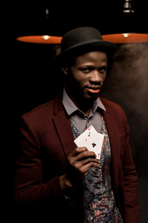 handsome elegant african american man with playing cards in hands in dark room with lampsの写真素材