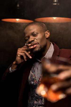 handsome luxury african american man smoking cigar and drinking whiskey in dark room with lampsの写真素材