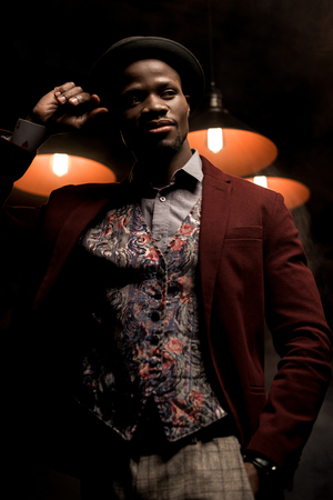 handsome african american man in hat posing in dark room with lampsの写真素材