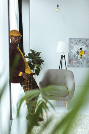 handsome stylish african american man pising in checkered pants, cap and jacket in modern officeの写真素材