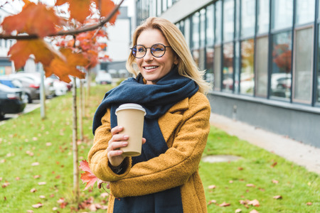 smiling attractive woman in yellow coat and scarf with coffee to go in autumn parkの写真素材
