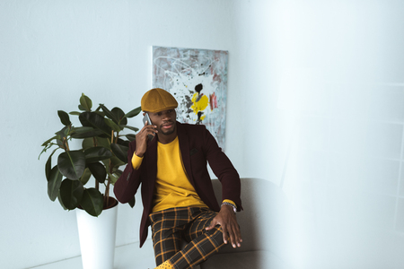 handsome fashionable african american businessman in cap and checkered pants talking on smartphone in modern officeの写真素材