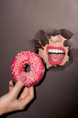 woman with pink lips trying to bite donut out of ragged hole in dark paperの写真素材