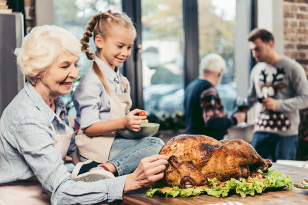 grandmother and granddaughter looking at turkey for holiday at kitchen while men talking blurred on backgroundの写真素材