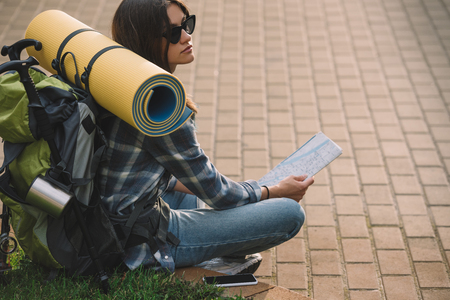 young female backpacker in sunglasses sitting on grass and holding mapの写真素材