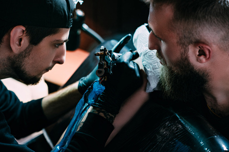 side view of tattoo artist in gloves with tattoo machine working on tattoo on shoulder in salonの写真素材