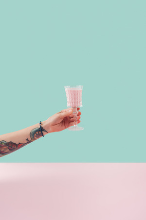 cropped view of female tattooed hand with glass of milkshake on pastel background