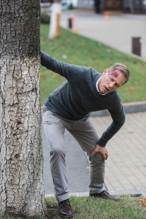 injured middle aged man with wound on head leaning on tree on the streetの写真素材