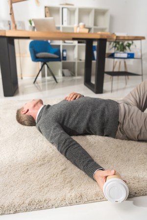 unconscious man lying on a floor in office and holding disposable coffee cup in handの写真素材