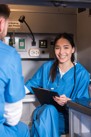 smiling Asian paramedic sitting in ambulance and looking at camera
