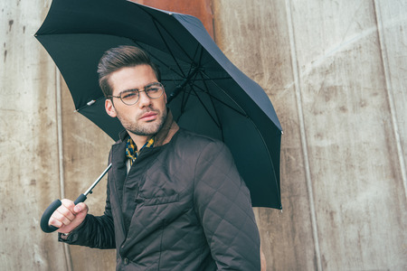 handsome stylish young man in eyeglasses holding umbrella and looking awayの写真素材