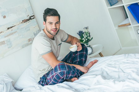 handsome smiling young man in pajamas holding cup of coffee and looking at camera while sitting on bedの写真素材