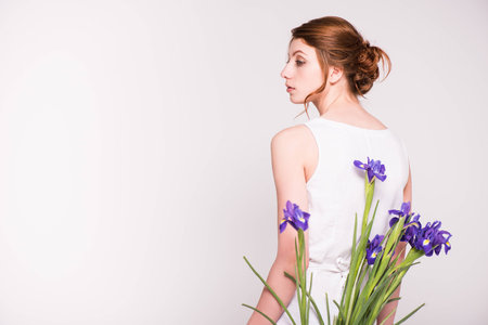 back view of gorgeous young woman in white dress holding iris flowers and looking away isolated on greyの写真素材