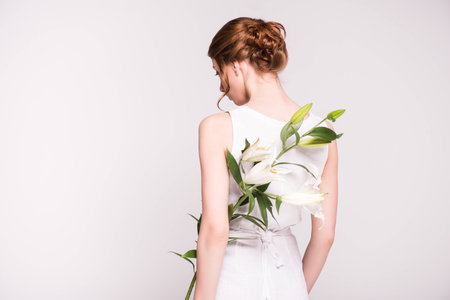 back view of young woman in white dress holding beautiful lily flowers isolated on greyの写真素材