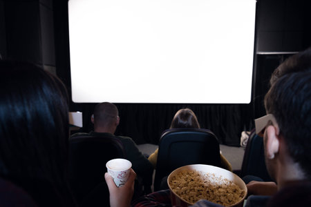 back view of friends with popcorn and drink sitting in cinema with white blank screen