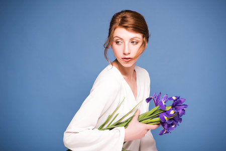 attractive young woman holding flowers and looking away isolated on blue